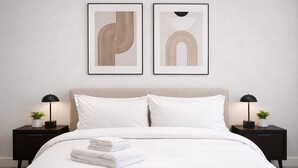 Una cama bien hecha con sábanas blancas y dos almohadas, flanqueada por dos mesitas de noche con lámparas y pequeñas macetas. Dos obras de arte abstracto decoran la pared sobre la cama.
