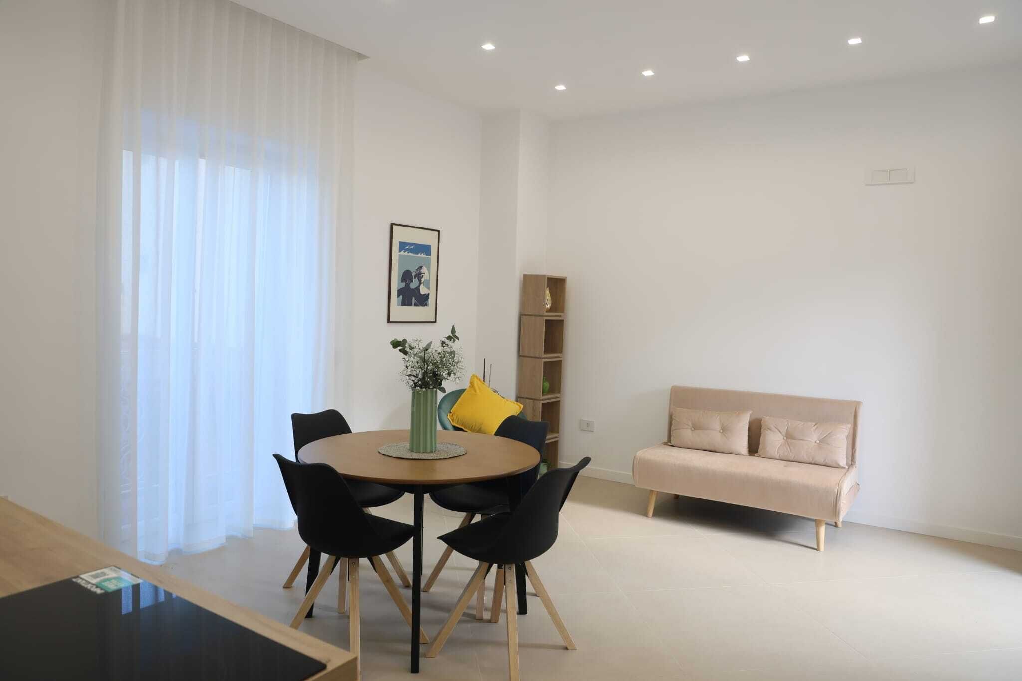 A modern living room with a round dining table, black chairs, a beige sofa, and a bookshelf.