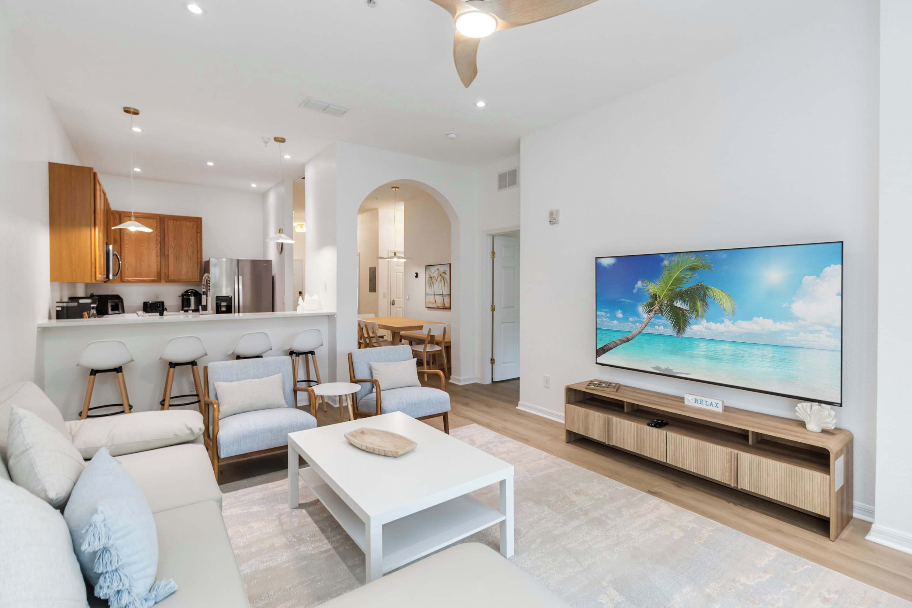 A modern living room with a large flat-screen TV, a sofa, a coffee table, and a kitchen area with wooden cabinets and a dining set.