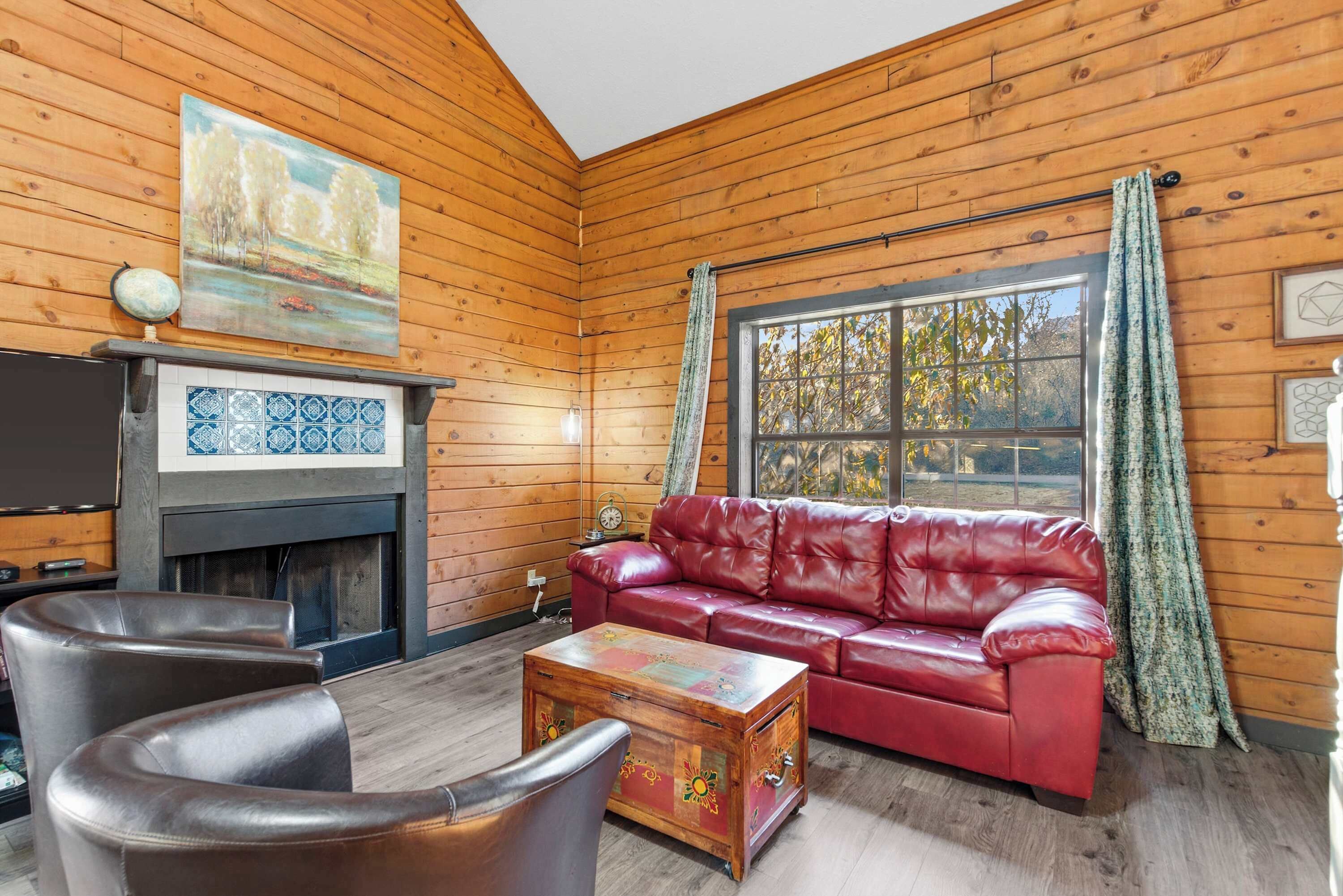 A cozy room with wooden walls, a red leather sofa, a fireplace with blue tiles, and a large window with curtains.