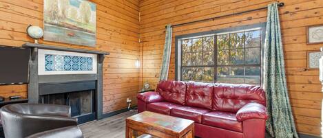A cozy room with wooden walls, a red leather sofa, a fireplace with blue tiles, and a large window with curtains.