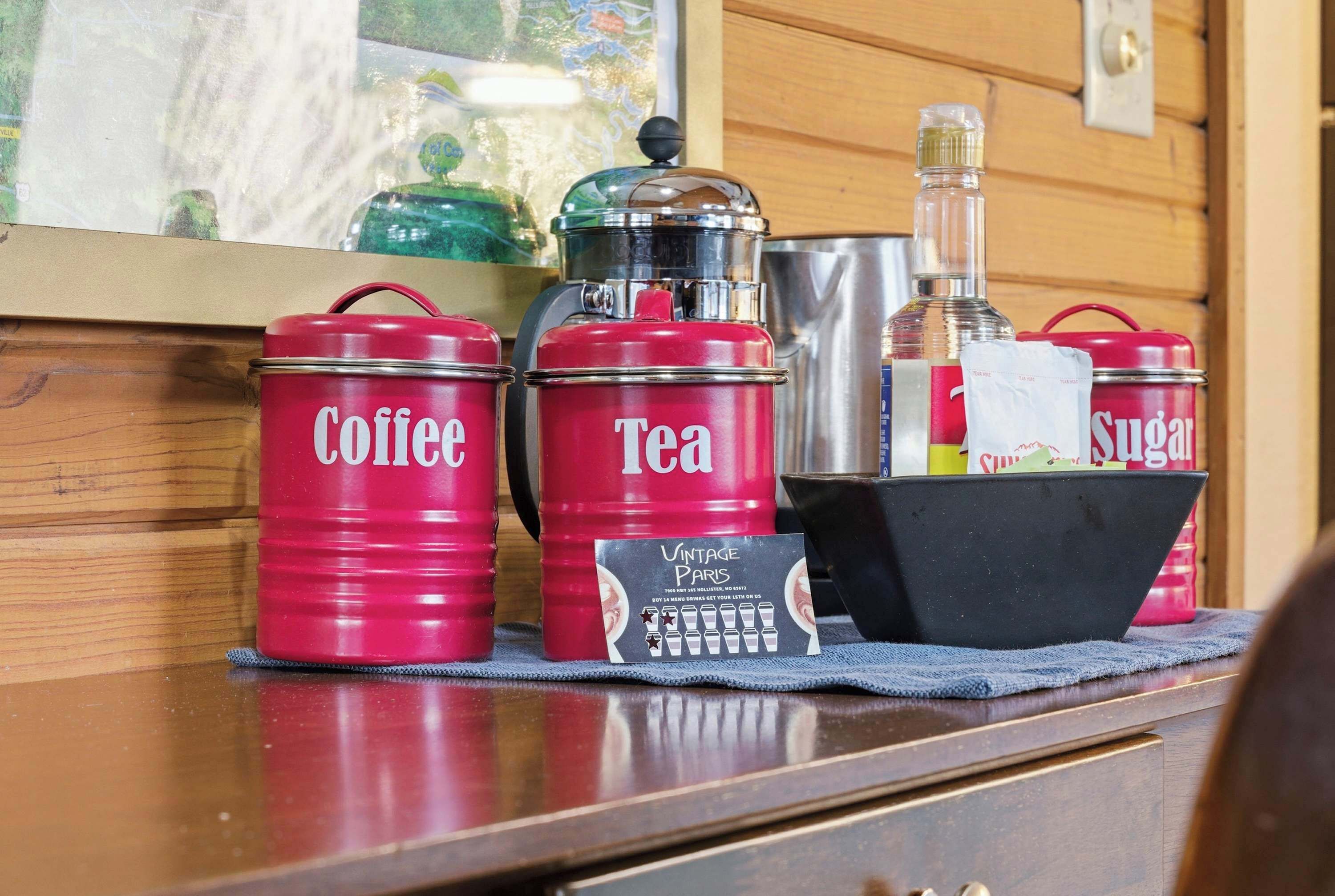 Red containers labeled "Coffee" and "Tea" on a wooden surface, with a card and a black basket in front.