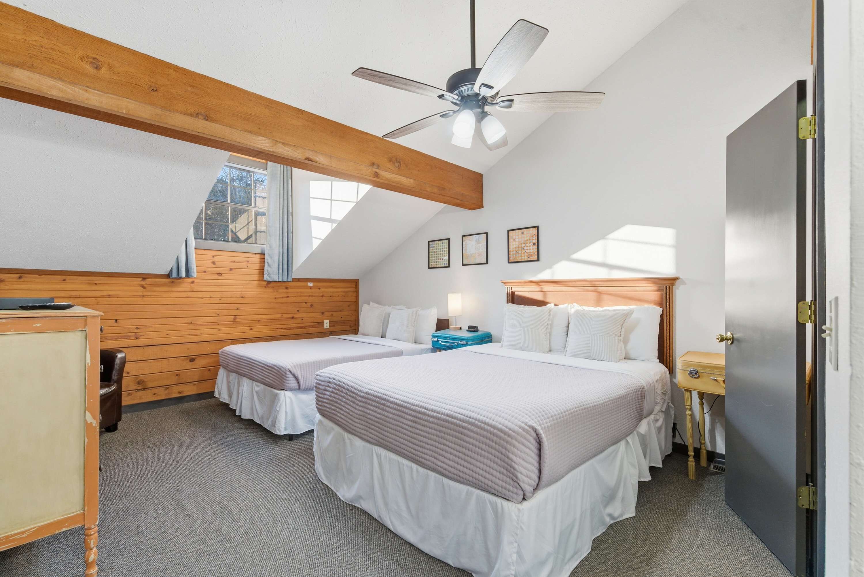 A bedroom with two beds, a ceiling fan, wooden beams, a window, and a door.