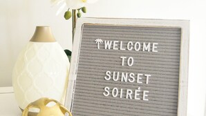 A decorative sign with "WELCOME TO SUNSET SOIRÉE" text, a white vase, and a golden decorative object.