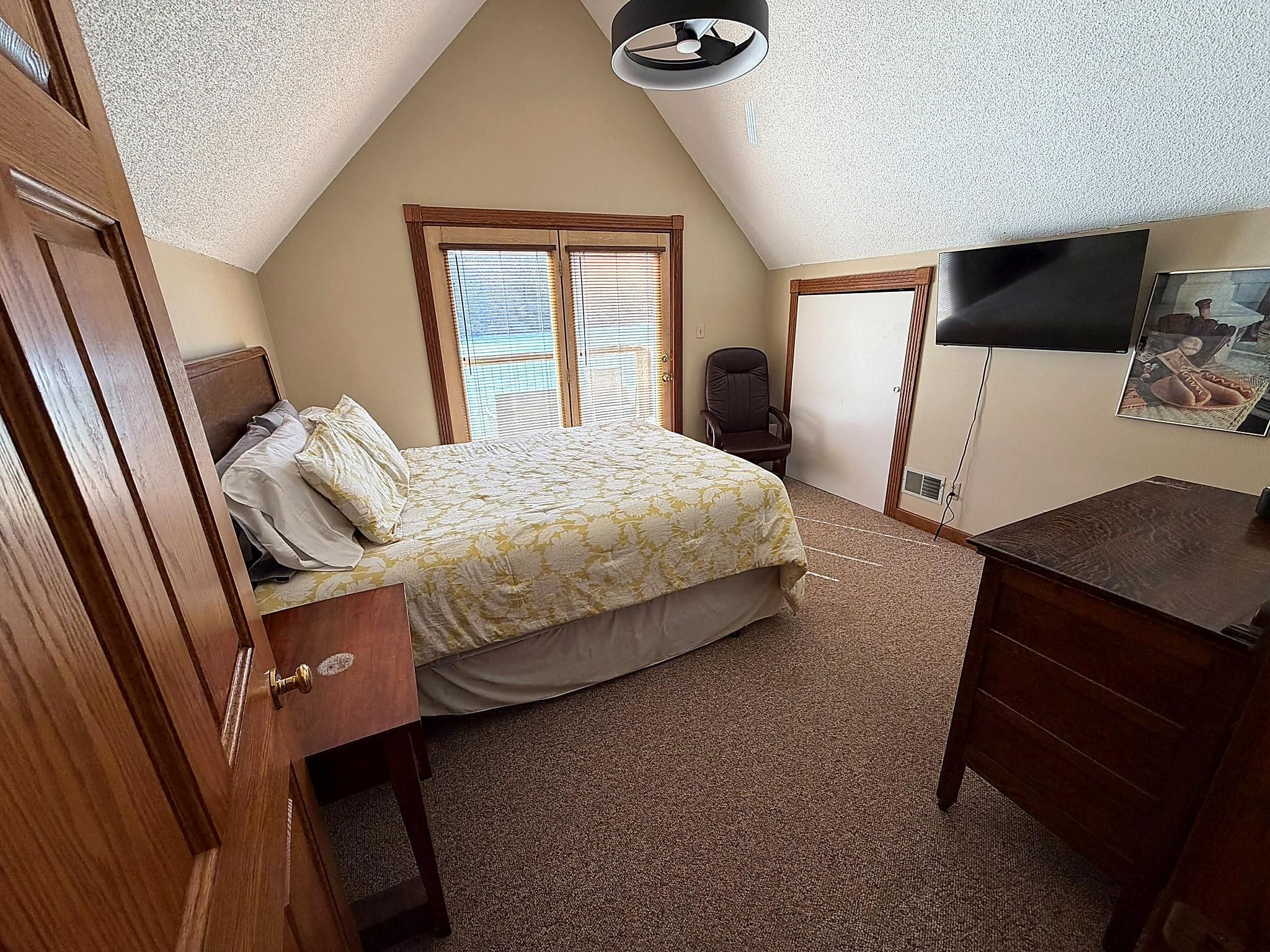 A bedroom with a bed, a desk, a chair, a television, and a window with blinds.