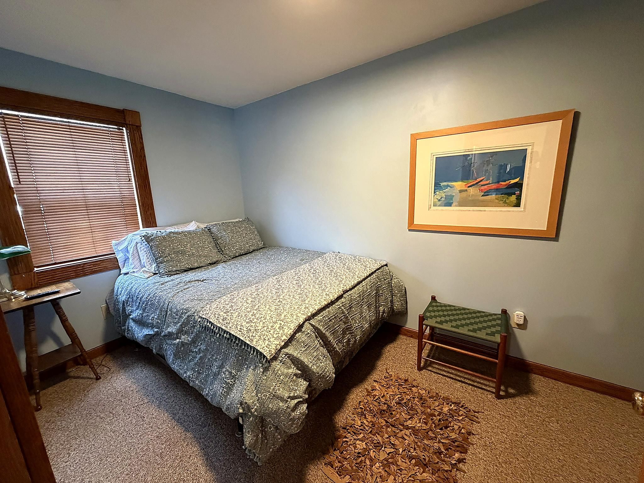 Quarto com cama, mesinha, cadeira, quadro na parede e janela com persianas.