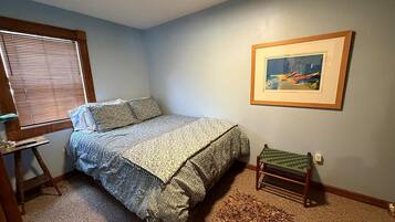 Quarto com cama, mesinha, cadeira, quadro na parede e janela com persianas.