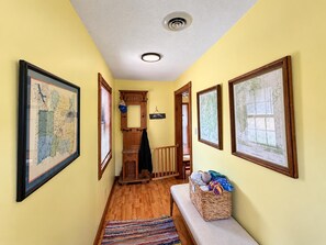 A hallway with wooden flooring, a bench with a basket, and framed maps on the walls.