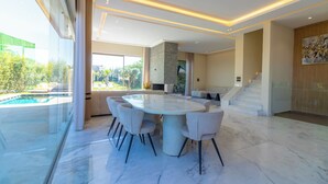 Un'elegante zona pranzo con tavolo in marmo e sedie, una scala e una piscina visibile attraverso ampie finestre.