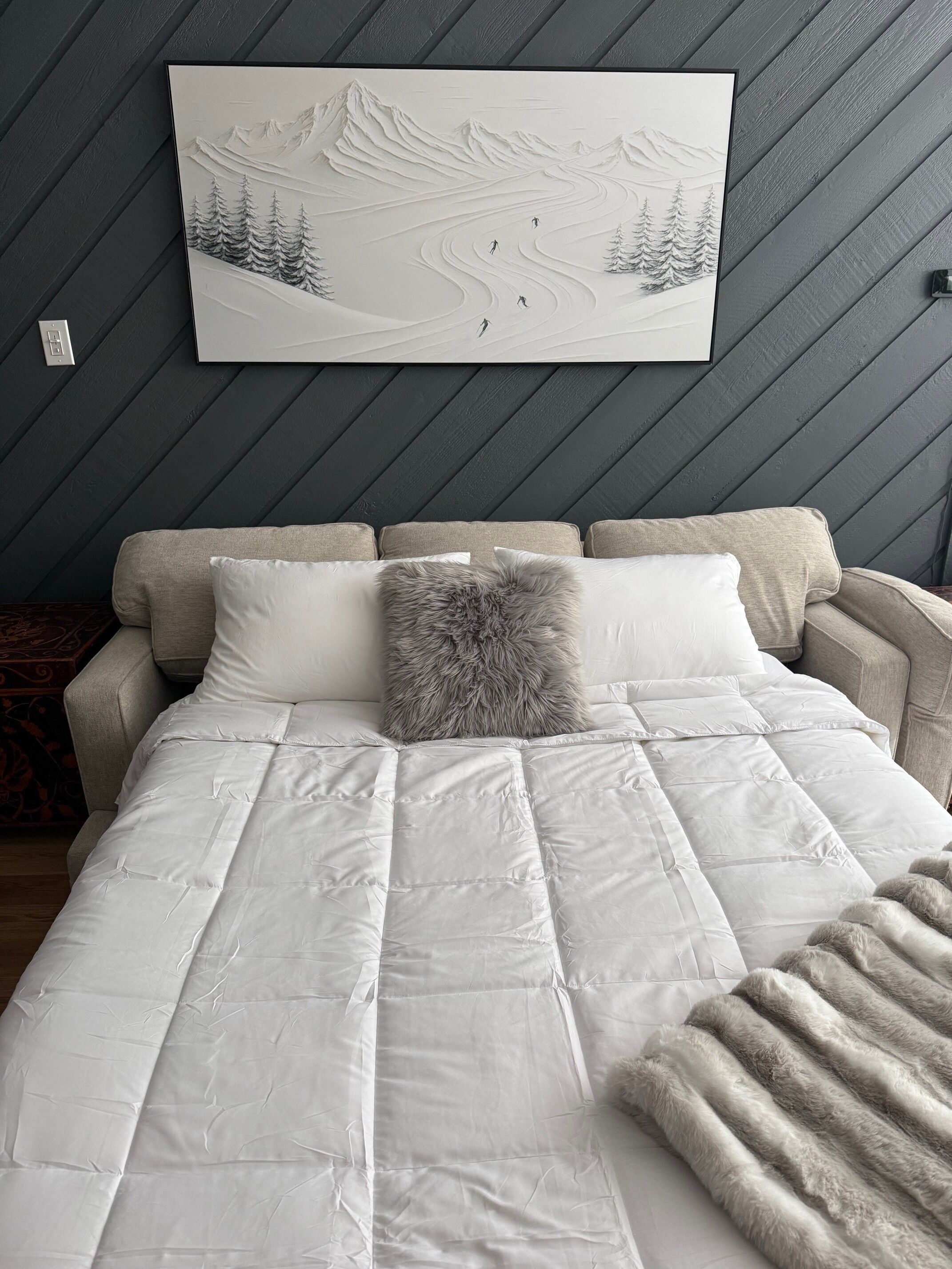 A bedroom with a bed, a grey furry throw, and a mountain landscape painting on the wall.