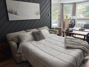 A modern bedroom with a sofa bed, a large painting of a mountain landscape, a dining area with a table and chairs, and a view of the outdoors through a window.