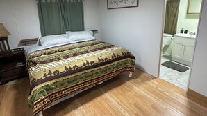 Un dormitorio con cama, mesita de noche, lámpara, cortina verde, cartel enmarcado y baño visible a través de una puerta abierta.