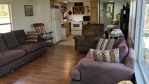 Una sala de estar con un sofá, una silla y una mesa de comedor. Se observa una cocina al fondo con estufa y armarios.