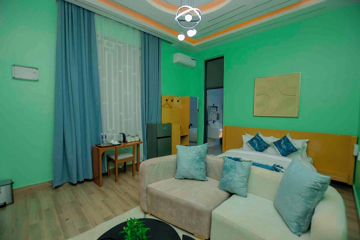 Una habitación de hotel moderna con una pared verde, un sofá color crema, una mesita de madera pequeña y una cama con ropa de cama azul y blanca.