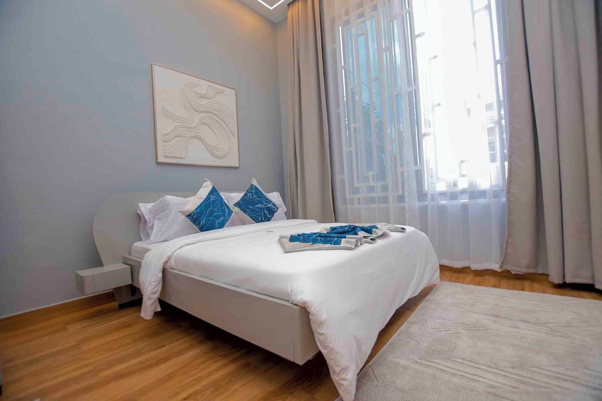 Un dormitorio moderno con cama, mesita de noche, una obra de arte en la pared y un gran ventanal con cortinas.