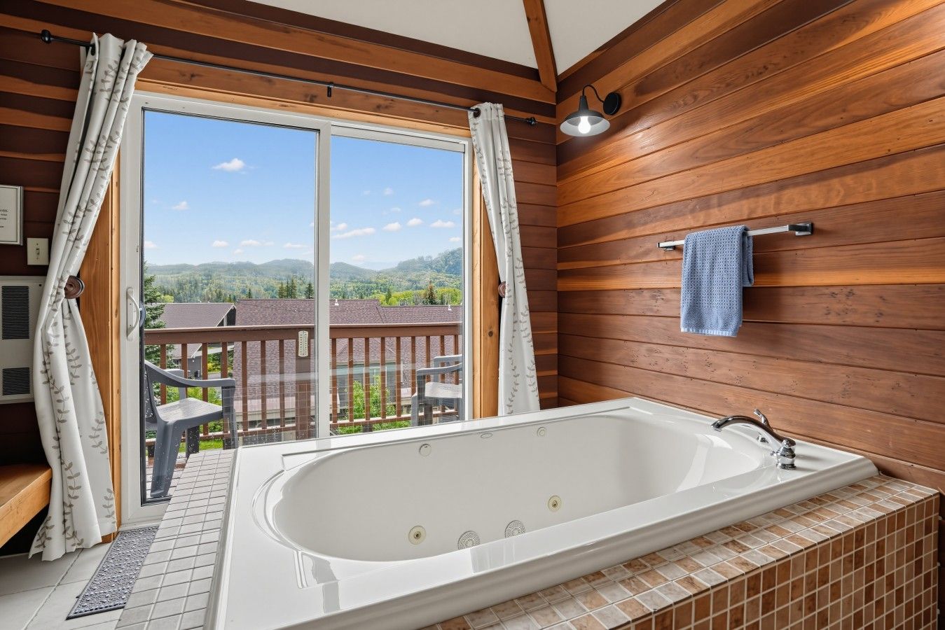 A bathroom with a large bathtub, wooden walls, a view of a balcony with a chair, and a towel hanging on the wall.