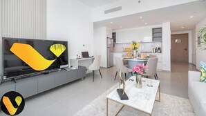 Un salon moderne doté d’un téléviseur à écran plat, d’un coin repas avec une table et des chaises, et d’une cuisine en arrière-plan.