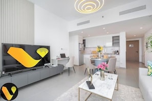A modern living room with a flat-screen TV, a dining area with a table and chairs, and a kitchen in the background. - Vogue | Downtown with Direct Access to Dubai Mall (Dubai)