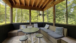 Ein überdachter Außenbereich mit Holzboden, großen Fenstern und einem gewebten Sofa mit Kissen.
