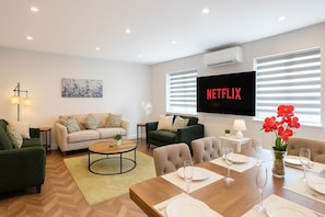 Een moderne woonkamer met een flatscreen-tv waarop 'NETFLIX' wordt weergegeven, een eethoek met een houten tafel gedekt voor vier personen, en twee banken.