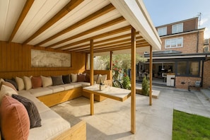 Un espace extérieur aménagé, abrité, avec un canapé et une table, un plafond en bois et une vue sur un immeuble d’habitation en arrière-plan.
