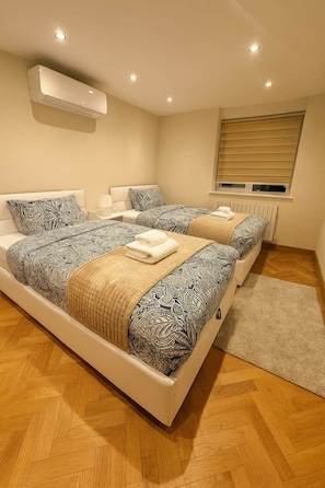 A modern bedroom with two beds, a built-in air conditioner, a window with blinds, and a herringbone wooden floor. - Beautiful Luxury home London | 10 bed 7 Bathroom (London)