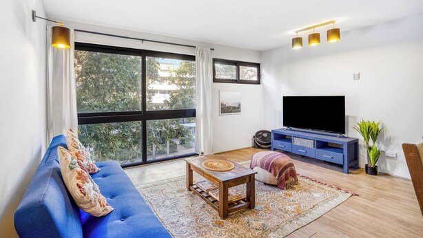 Un salon moderne avec un canapé bleu, une table basse, un téléviseur à écran plat et une grande fenêtre donnant sur des arbres.