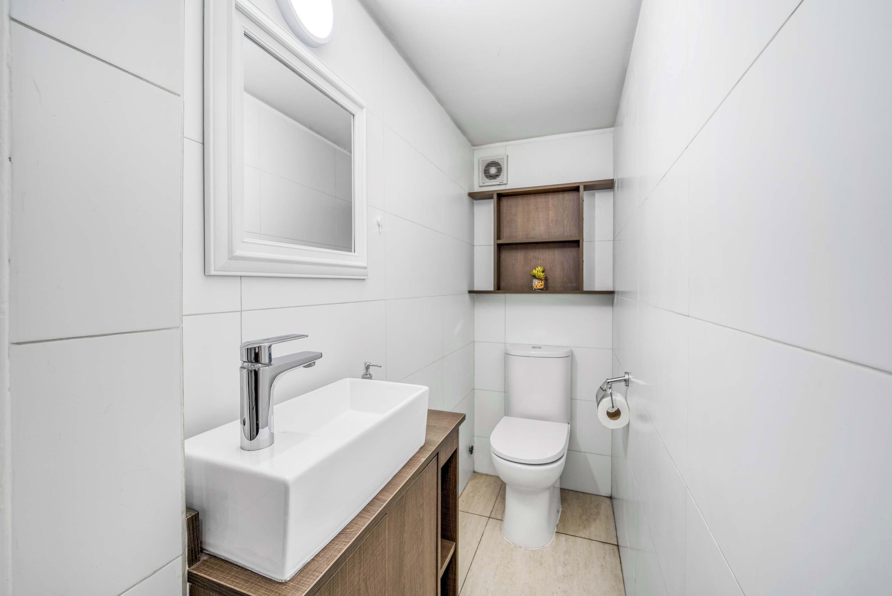 Ein modernes Badezimmer mit einem weißen Waschbecken, einem Spiegel, einer Toilette und einem Regal mit einer kleinen Dekoration.