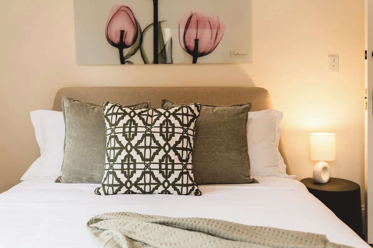 Een net opgemaakt bed met gestreepte kussens, een schilderij van roze bloemen aan de muur en een bedlampje op een klein tafeltje.
