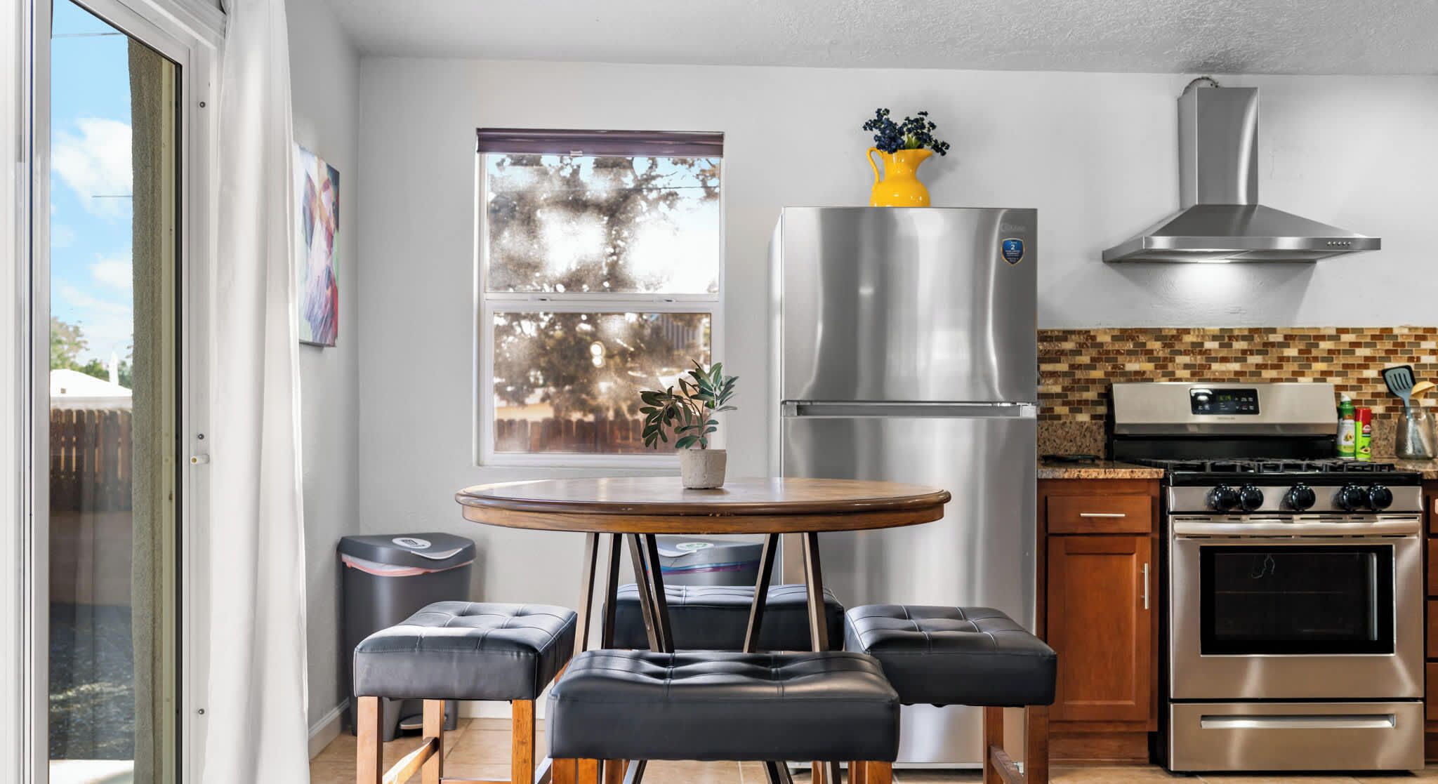 A modern kitchen with a stainless steel refrigerator, oven, and stove, a round wooden table with four black leather stools, and a window with a view of trees.