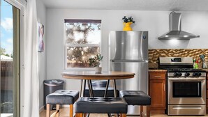 Una cocina moderna con un refrigerador, horno y estufa de acero inoxidable, una mesa redonda de madera con cuatro taburetes de cuero negro, y una ventana con vista a árboles.