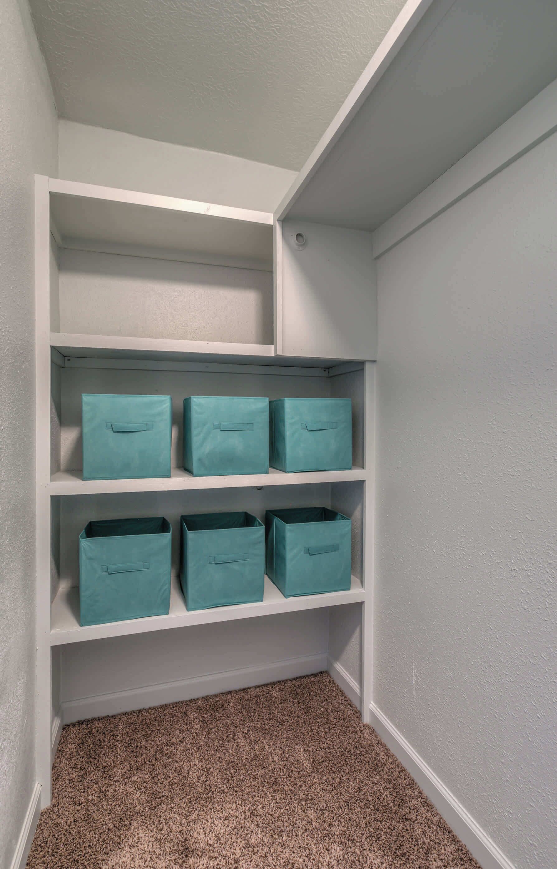 A closet with white walls and a carpeted floor, containing four teal storage bins on the shelves.