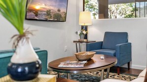 Una sala de estar con un sillón azul, una mesa redonda de centro con un tazón y un control remoto, una lámpara y un cuadro en la pared.