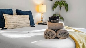 Cama bien hecha con toallas enrolladas y una toalla con la palabra 'Makeup', una lámpara y una planta en una habitación de hotel.