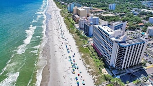 Een kuststadsgezicht met een lang strand, langs de kust gebouwd met verschillende gebouwen, waaronder een opvallend hotel met meerdere verdiepingen en balkons.