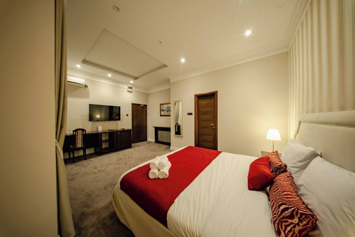 A hotel room with a neatly made bed, a desk with a chair, a television, and a fireplace.