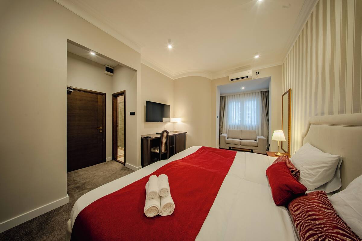 A hotel room with a large bed, a desk, a chair, a TV, and an air conditioning unit.