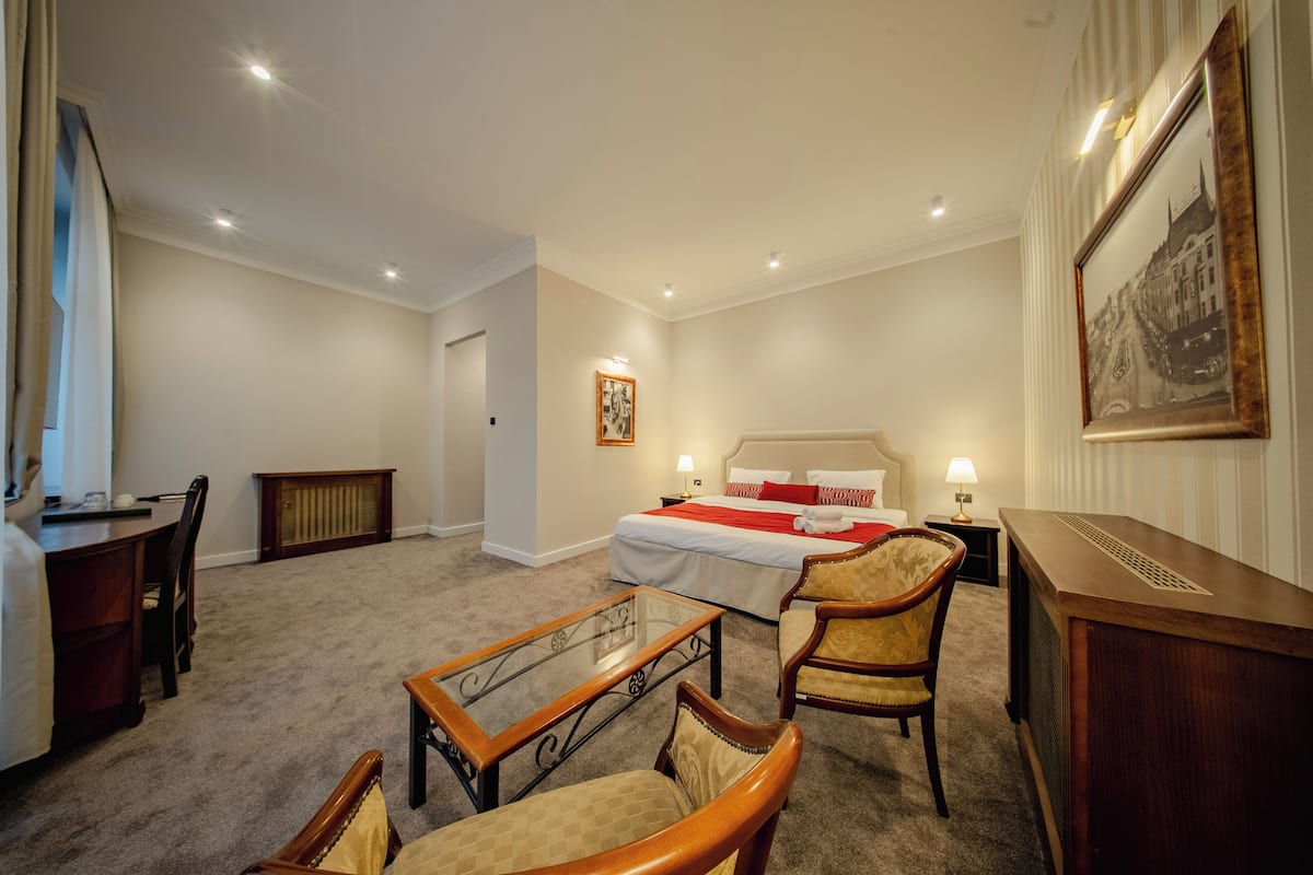 A hotel room with a bed, a desk, a chair, a small table, and a radiator.