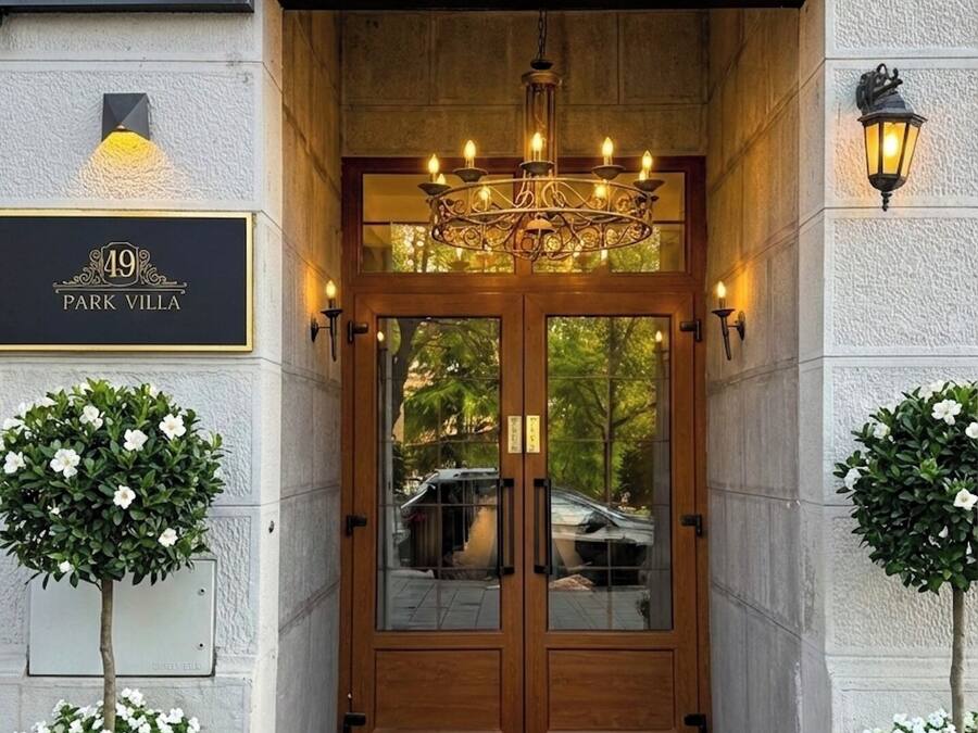 Entrada de hotel com tapete vermelho, porta dupla de madeira com painéis de vidro, um candelabro e duas plantas em vasos.