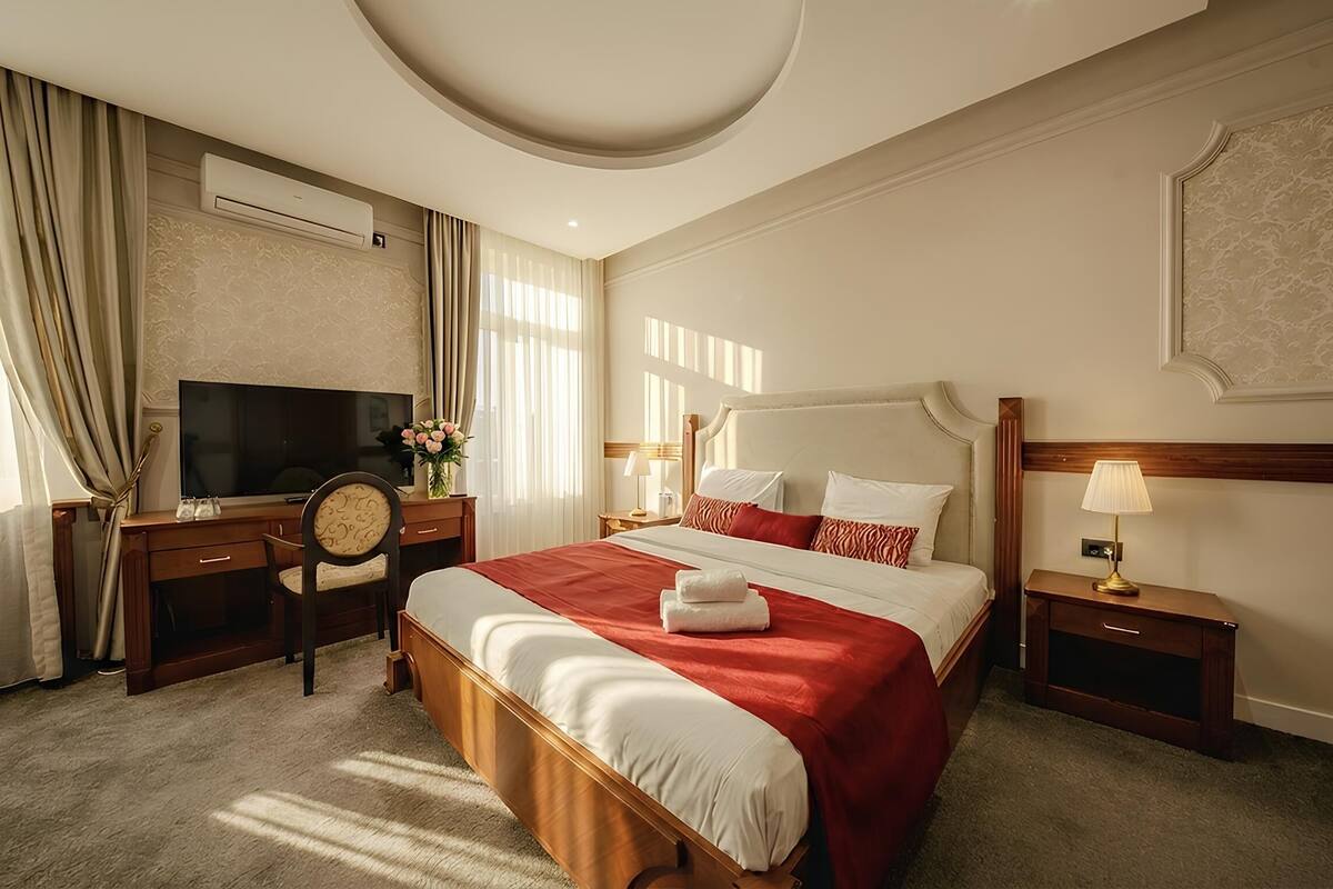 A hotel room with a large bed, a desk with a TV, a chair, and a nightstand with a lamp.