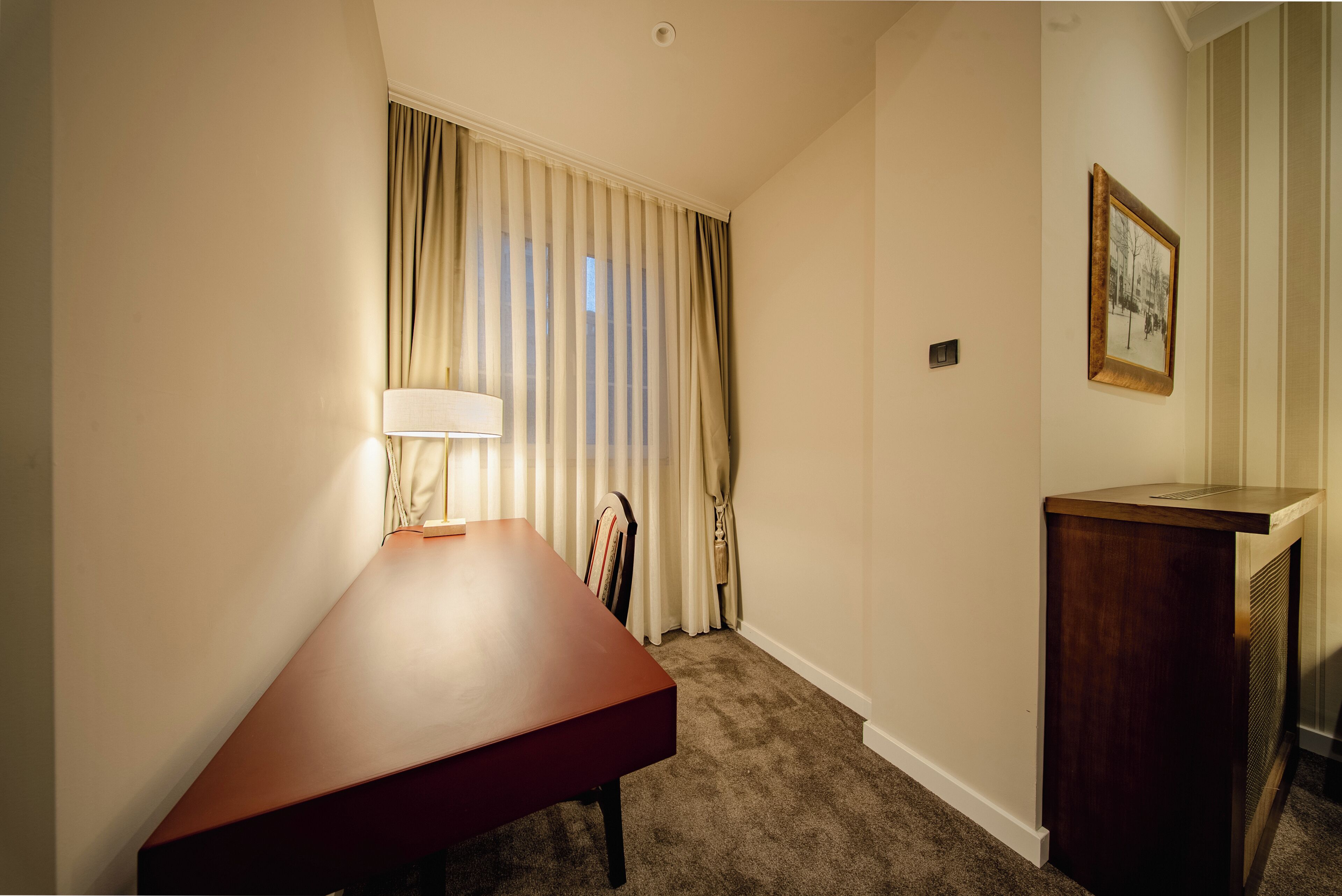 A hotel room with a wooden desk, a lamp, a framed picture on the wall, and a window with curtains.