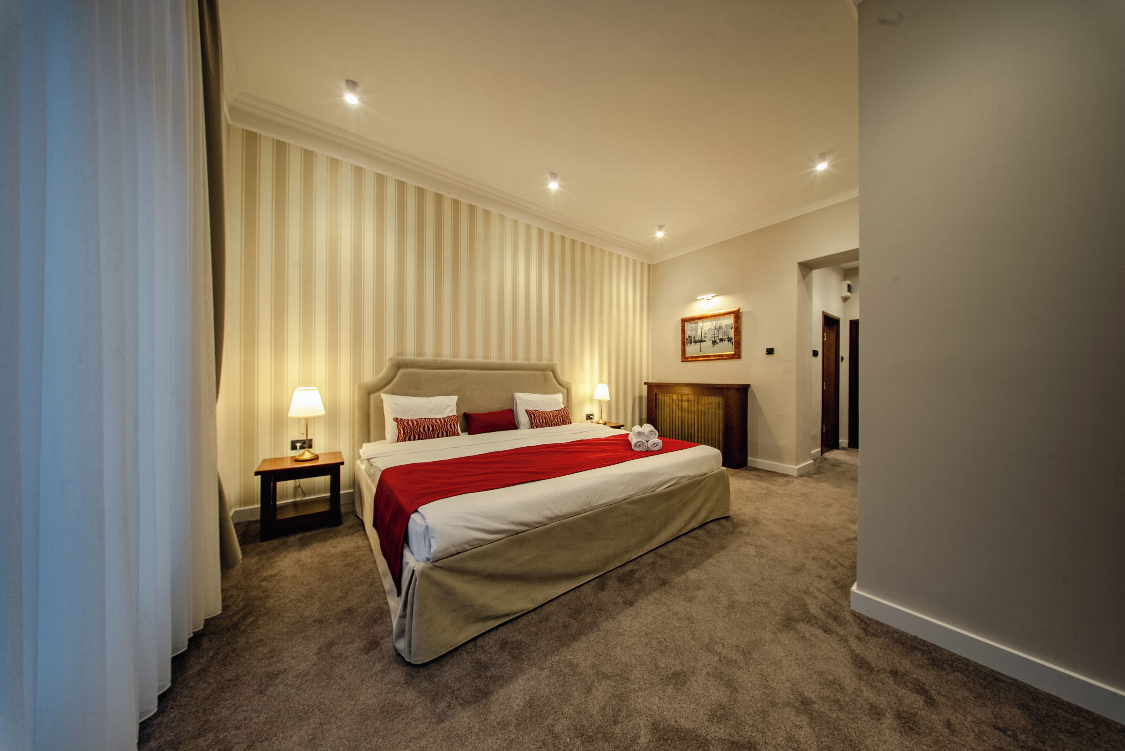A hotel room with a large bed, a nightstand, a lamp, a framed picture on the wall, and a door leading to another room.