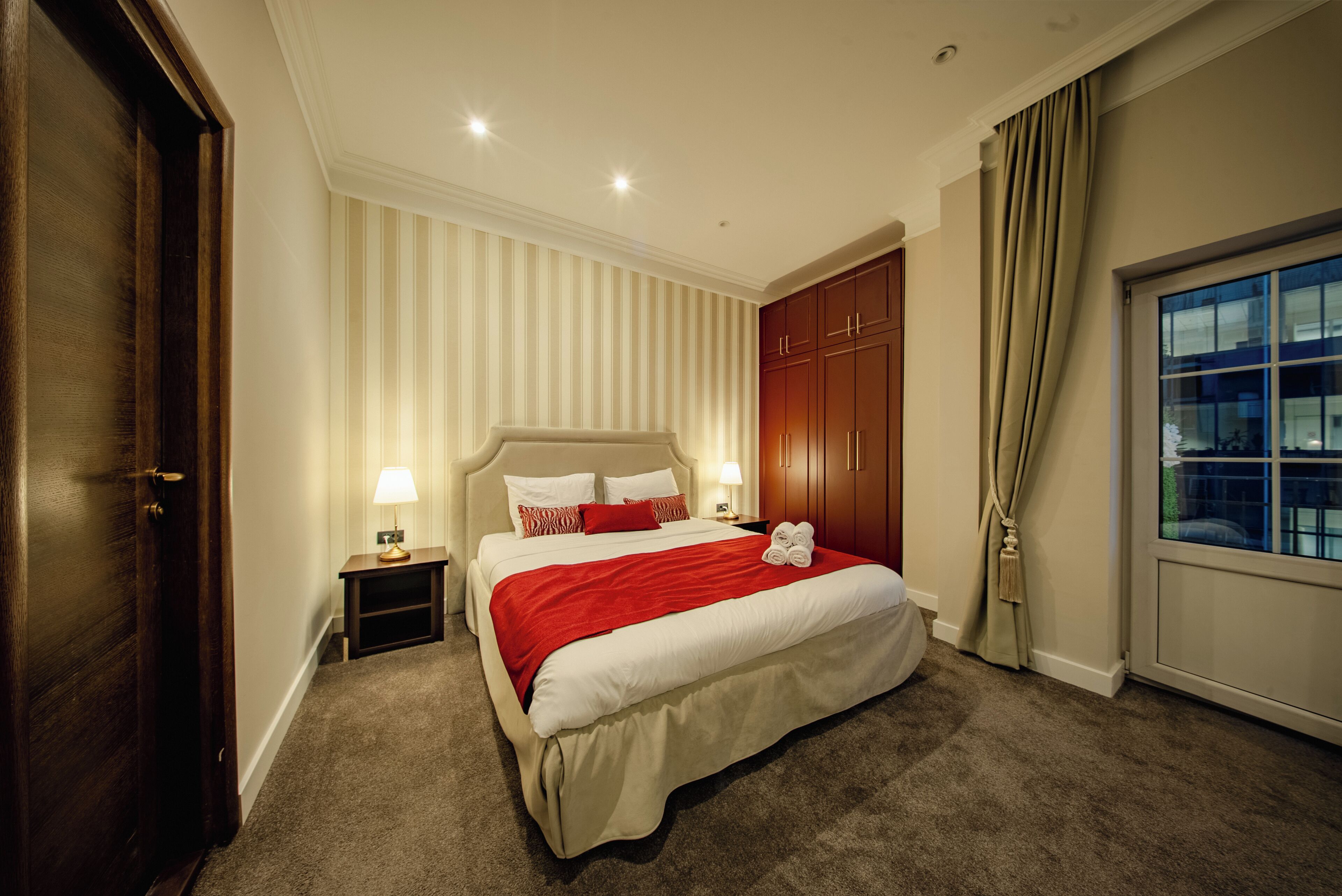 A hotel room with a large bed, a wooden wardrobe, a nightstand with lamps, and a window with curtains.