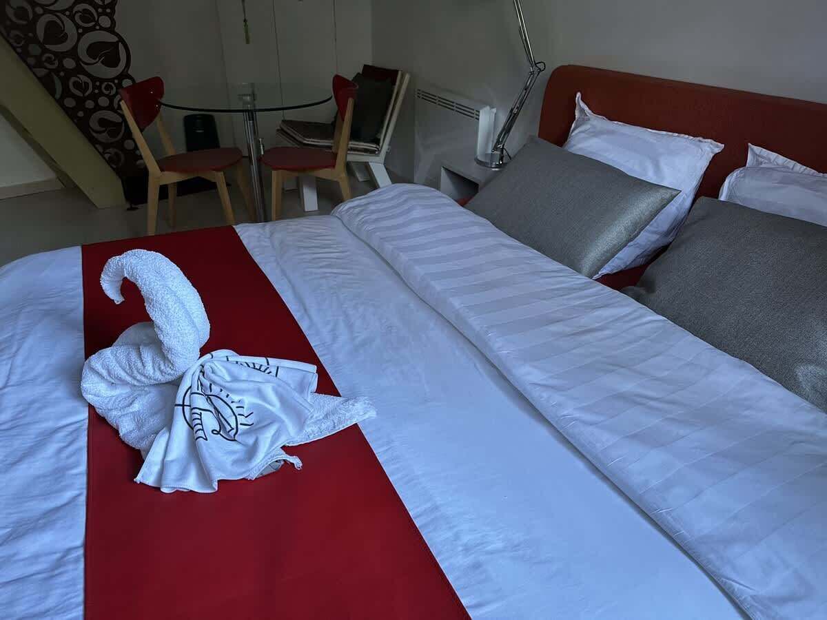 En pænt redt seng med et rødt farveaccent, et hvidt håndklæde med svane-design og en grå pude.