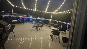 Una terrazza all'aperto con luci a catena, tavoli e sedie, con vista su un panorama urbano notturno.