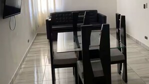 En moderne stue med en sort sofa, et glasbord med stole, et fladskærms-tv på væggen, en loftventilator og et vindue med gardiner.