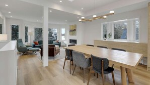 Ein modernes Wohnzimmer mit einem großen Esstisch aus Holz, bequemen Sitzgelegenheiten und großen Fenstern mit Blick ins Freie.