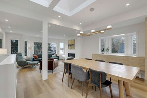 Ein modernes Wohnzimmer mit einem großen Esstisch aus Holz, bequemen Sitzgelegenheiten und großen Fenstern mit Blick ins Freie.
