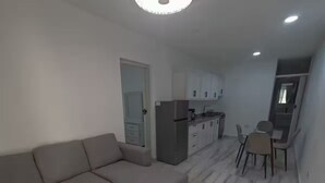 Una sala moderna con un sofá seccional gris, un comedor con mesa y sillas, y una cocina con gabinetes blancos y un refrigerador.