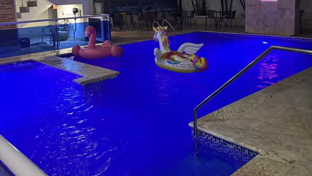 Una piscina con tonalidades azules, rodeada por un edificio moderno que incluye una escalera y un mural con una escena de playa.
